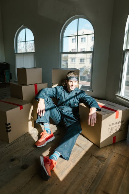A young man with shoulder-length hair, wearing a patterned headband, a blue work jumpsuit, and bright orange sports shoes, is sitting on a wooden floor inside a well-lit room with large arched windows. He is surrounded by several closed cardboard boxes, some with red and black packing tape, indicating they are prepared for transportation. The natural light from the windows casts shadows on the floor, highlighting the boxes and the man's relaxed posture. This scene appears to be part of a home relocation or packing process, with the boxes likely containing household items in preparation for a household move, supported by the presence of the boxes and the indoor environment. The interior space is minimalistic with light-colored walls and wooden flooring, typical of a residential property. This image is associated with professional removals services, focusing on packing and moving logistics, as seen on the webpage about narrow-street removals in W6, Ravenscourt Park, provided by manwithavanravenscourtpark.co.uk.