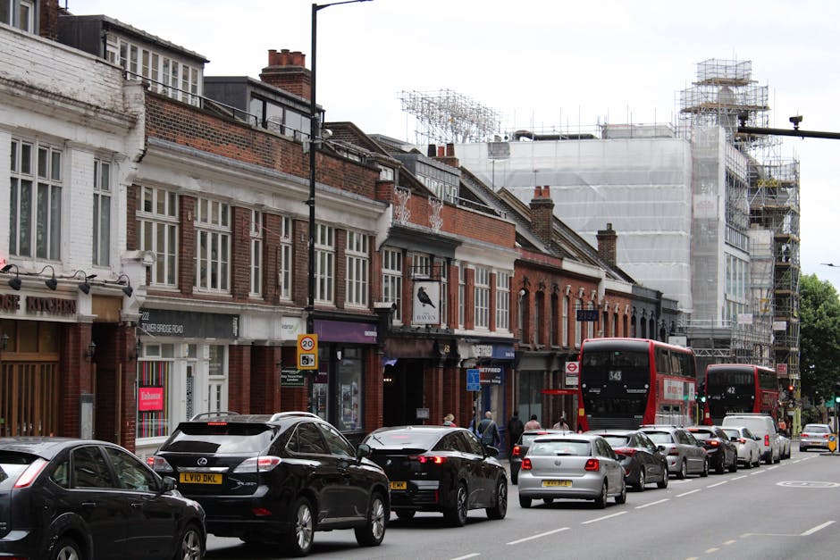 A street scene in Brackenbury Village with a row of brick and white-painted terraced houses, some of which are under renovation with scaffolding around the upper floors. The ground floor shop fronts include a furniture store and other retail outlets. Parked along the curb are several vehicles, including black, silver, and grey cars, with two red double-decker buses passing by. Pedestrians are walking on the sidewalk, some near the storefronts. In the foreground, there is a city street with vehicle loading and unloading activities consistent with home removals or furniture transport, and two vans from a removals company are visible near the street, likely involved in a home relocation process. The scene is captured during daytime with natural lighting, highlighting the building details, parked cars, and ongoing construction work on the buildings.