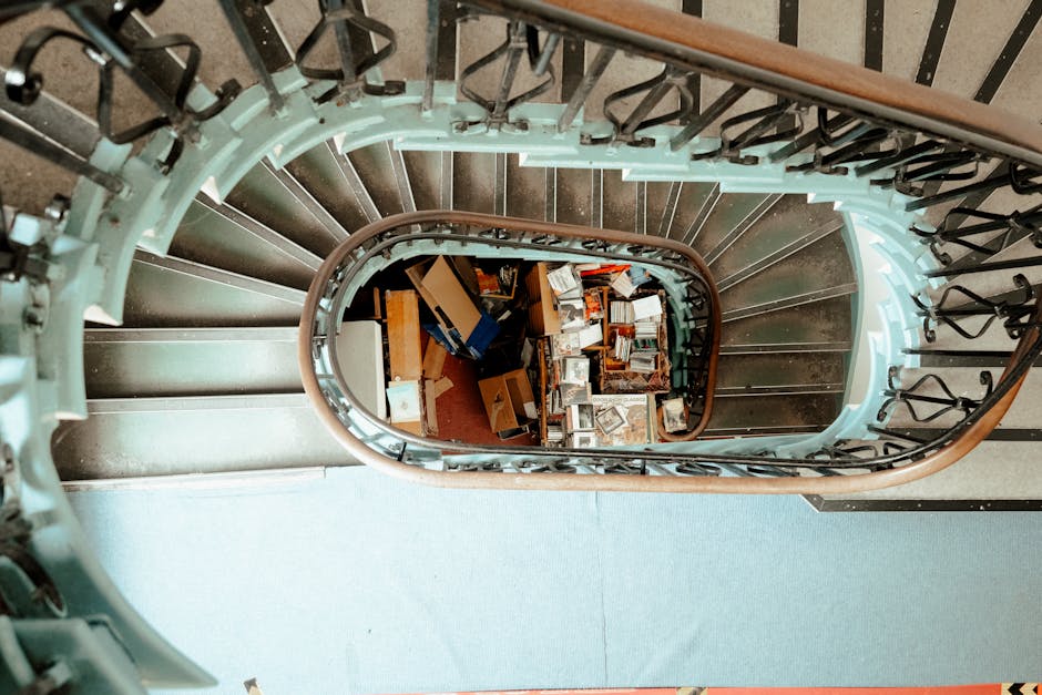 A view from the top of a spiral staircase inside a property, showing the staircase's black metal balustrade with decorative elements and a wooden handrail. The stair treads are dark-colored, with a light-colored riser. At the bottom of the staircase, there is a piled assortment of cardboard boxes, some open with visible contents such as books, small household items, and packaging materials. The boxes are of various sizes, placed on a red carpeted floor, which is partially visible. The surrounding environment includes a light-colored wall and a carpeted landing area at the top of the staircase. This scene captures the process of home relocation, with furniture and belongings prepared for transport as part of a removal or moving service by [COMPANY_NAME], illustrating the challenges of loading bulky items down narrow, tight stairs typical of residential properties on Glenthorne Rd. The image emphasizes the careful handling of packed boxes in the context of furniture transport and packing during house removals.