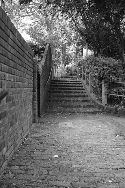 A black-and-white photograph of a narrow, inclined outdoor staircase made of stone or concrete, situated between a brick wall on the left and a garden with bushes and trees on the right. The stairs lead upward through a shaded area with dappled light filtering through the foliage above. At the top of the staircase, the path continues with more greenery visible. This setting appears to be within a residential area, likely part of a house relocation or removal process, with the staircase potentially used for moving bulky furniture from a property on Glenthorne Road, as referenced in the page title, towards a vehicle for transportation. The overall environment suggests a quiet, urban residential street, suitable for professional removals handling furniture transport up or down tight, inclined access points.