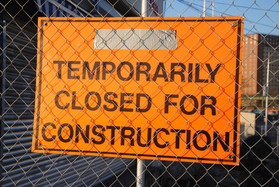 Close-up of an orange rectangular sign with black text reading 'TEMPORARILY CLOSED FOR CONSTRUCTION' affixed to a metal chain-link fence. The sign is in front of a construction site with visible metal panels, a partially constructed building, and a clear blue sky. The sign indicates that the area or premises is temporarily inaccessible due to ongoing construction work, with the fence serving as a barrier. The location appears to be in an urban setting, and the image is consistent with visual cues related to building or home relocation activities, which may involve coordination with construction zones as part of moving or packing logistics.
