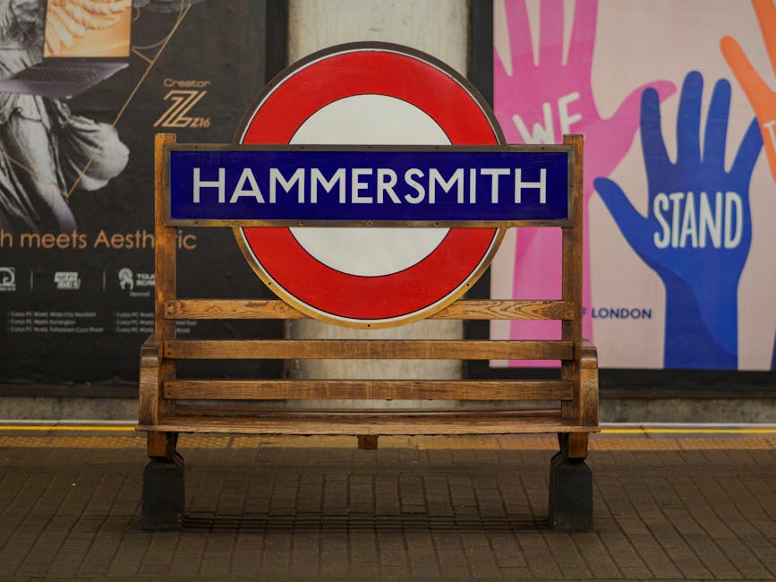 A wooden bench with horizontal slats sits on a tiled pavement in front of a large, circular London Underground sign that reads 'HAMMERSMITH' in white letters on a blue background, framed by a red and