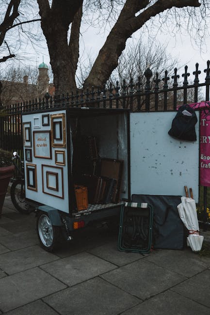 A small, mobile removal cart parked on a sidewalk near a black metal fence, with its side panel open revealing various framed pictures, cardboard boxes, and packing materials inside. The cart has a wh
