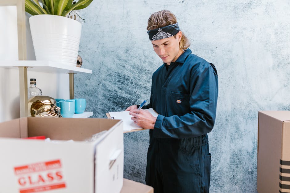 A young man with light skin, wearing a navy blue uniform and a black bandana with a white pattern, is inside a room with a textured grey wall. He is standing next to a white shelving unit that holds a large white ceramic pot with a plant, blue cups, and a clear glass bottle. In front of him are several cardboard boxes, one of which has red and white printing, likely used for packing and moving purposes. The man is writing on a clipboard, noting details relevant to a house or office relocation process. The lighting is natural, illuminating the scene clearly, suggesting a professional setting focused on packing, moving, or inventory checking as part of a furniture transport or home relocation service provided by [COMPANY_NAME]. This image represents careful planning, organization, and logistics involved in house removals or office moves, with an emphasis on packing materials and moving preparations.