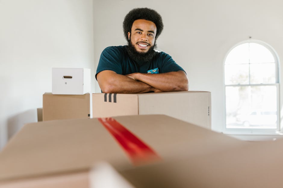 A smiling man with a beard and curly hair standing behind a stack of cardboard moving boxes inside a well-lit room with plain white walls and a large arched window in the background. The boxes vary in size, with one small white box on top of a larger brown one, and several others around him. The boxes are sealed with packing tape, and some appear to contain household or office items, indicative of a home relocation process. The room features natural daylight coming through the window, illuminating the scene clearly. The man is dressed in a dark blue t-shirt and has his arms crossed on top of the boxes, suggesting that he is either preparing for or involved in a furniture transport or packing and moving activity. This setting visualises professional removals and business relocation services, aligned with house removals and furniture transport assistance provided by [COMPANY_NAME].