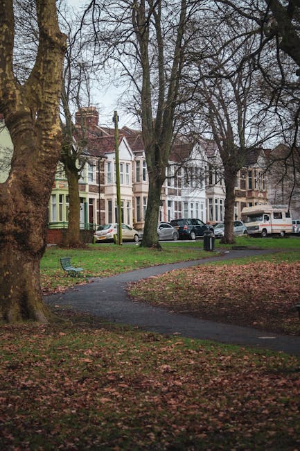 A residential street scene showing a paved pathway winding through a park area with leafless trees and fallen autumn leaves covering the ground. In the background, there are several multi-storey Victorian-style houses with bay windows and brick facades, alongside parked cars including sedans and a camper van. The park area features a bench on the grass near the pathway. The overall setting appears to be during overcast weather, with soft natural light illuminating the scene, providing a calm and neutral environment consistent with a typical UK neighbourhood prepared for home relocation or moving services. This image aligns with the concept of house removals, highlighting the exterior environment where packing and furniture transport logistics may occur, as seen in professional moving practices like those at manwithavanravenscourtpark.co.uk.