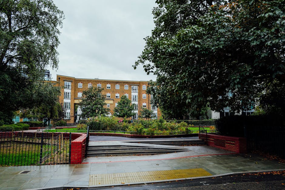 The image shows the exterior of a residential property with an entrance area leading to a garden or courtyard. In the foreground, there is a paved sidewalk with a tactile paving strip for accessibility, bordered by red brick walls and black metal railings. Wooden steps lead up to the garden, which is enclosed by additional fencing; some steps appear to be partially lifted or under maintenance. The garden features neatly maintained flowerbeds, shrubbery, and small trees, with a large residential building visible in the background. The building has multiple windows, some with white frames, and appears to be an apartment complex designed in a modern, brick-and-mortar style. The overall scene is captured during daylight hours, with overcast sky and diffused natural light, depicting a typical urban setting that may be involved in a house or apartment move process, supported by logistics specialists like [COMPANY_NAME] for furniture transport and packing during home relocation in the W6 area near Ravenscourt Park.