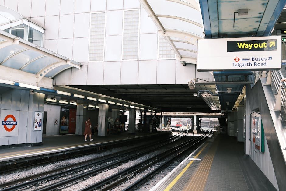 Exterior view of Hammersmith station showing train tracks in the foreground and the station entrance on the left, with a woman walking on the pavement near the platform. A sign displaying directions f