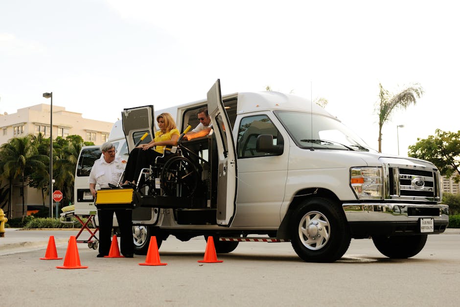 A white moving van parked on a street near residential buildings, with its side door open revealing a woman in a yellow top seated in a wheelchair being assisted by a man inside the vehicle, possibly preparing for a home relocation. A mover dressed in a white shirt stands outside, guiding the process, while orange traffic cones are positioned around the van to secure the loading area. The van is equipped with ramps and straps for furniture transport, and the scene is set during daylight with trees and a modern apartment complex visible in the background. This image illustrates the loading process involved in professional removals and furniture transport, emphasizing accessibility and careful handling during residential moves, as provided by [COMPANY_NAME] in W6.