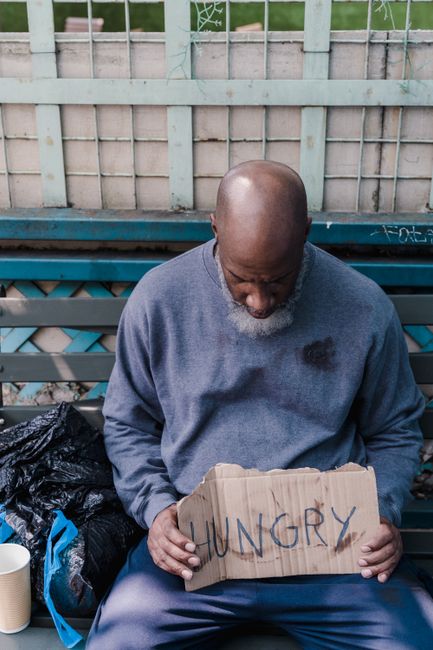A middle-aged man with a bald head and grey beard, dressed in a grey sweatshirt with visible stains, is sitting on a blue park bench outdoors. He is holding a cardboard sign with the word 'HUNGRY' written in large letters, and appears to be in a state of distress or concern. Behind him, there is a wooden fence with some graffiti and greenery peeking through. To his left, there are black plastic bags and a paper cup on the bench, indicating a setting associated with homelessness or hardship. The image captures a moment of vulnerability, which can relate to the emotional aspect of house removals and the importance of careful, compassionate moving services, as offered by [COMPANY_NAME], especially when dealing with complex or sensitive situations involving clients.